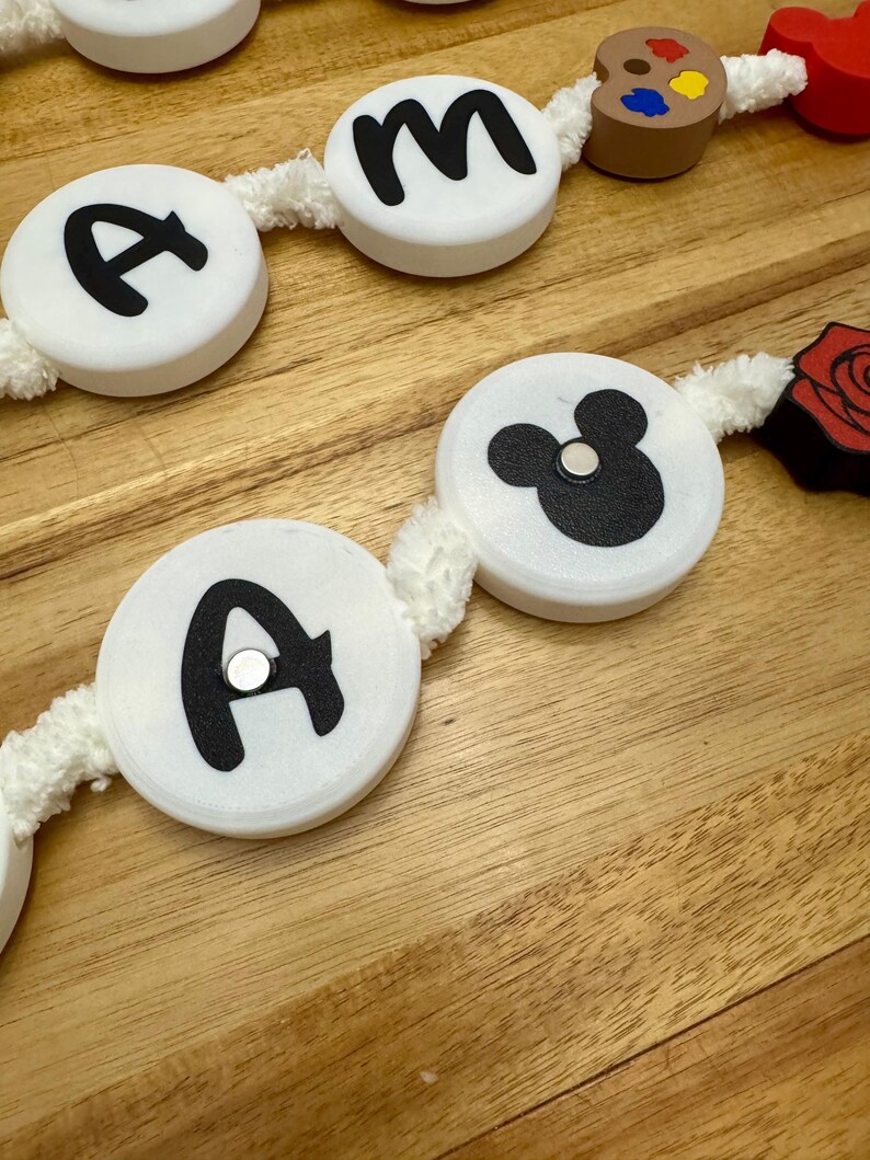 May include: A decorative garland featuring white circular beads with black letters and a Mickey Mouse silhouette. Additional elements include a wooden artist's palette and a red rose. The garland is strung on a white, fuzzy cord.