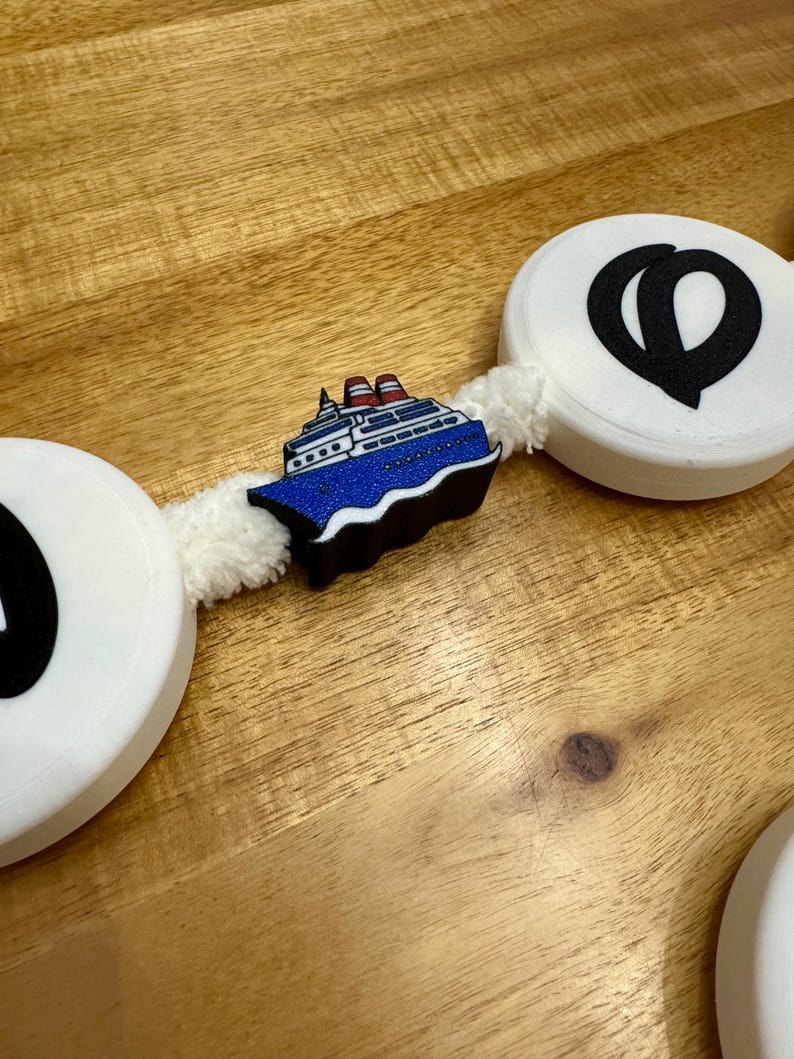 May include: A decorative garland featuring white circular elements with black graphic designs and a blue cruise ship charm. The garland is connected by a white, fluffy material, creating a nautical theme.