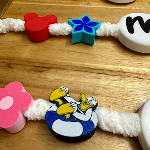 May include: A decorative garland featuring various shapes and characters, including a red Mickey Mouse head, a blue star, and a Donald Duck figure. The garland is strung on a white, fluffy cord and includes pink and white flower shapes and lettered discs.