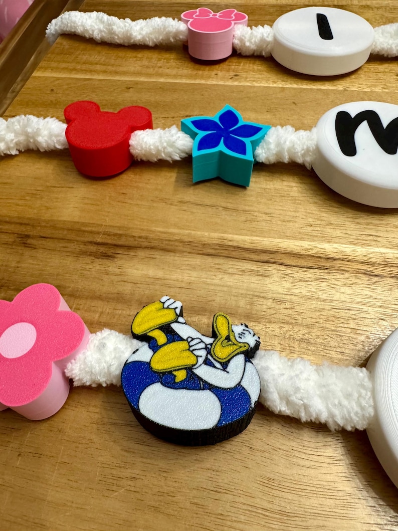 May include: A decorative garland featuring various shapes and characters, including a red Mickey Mouse head, a blue star, and a Donald Duck figure. The garland is strung on a white, fluffy cord and includes pink and white flower shapes and lettered discs.