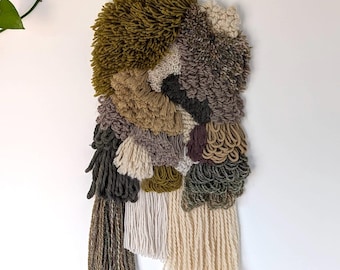 May include: A wall hanging made of yarn in shades of brown, green, and cream. The yarn is woven into a variety of textures, including loops, knots, and fringes. The wall hanging is a unique and modern piece of art.