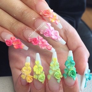 Könnte beinhalten: Nahaufnahme von langen, weißen, mandelförmigen Acryl-Nägeln mit 3D-Blumenmotiven. Die Blumen sind in Rosa-, Gelb-, Grün- und Blautönen gehalten und erzeugen einen Regenbogeneffekt. Die Nägel sind mit einer weißen Spitze und einer transparenten Basis verziert.