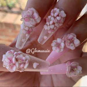 May include: Long, clear, almond-shaped acrylic nails with delicate pink 3D floral designs and gold accents. The nails are decorated with small pink and gold beads. The text "@Glamsusie" is visible on one of the nails.