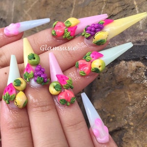 May include: Close-up of a hand with long, pointed nails painted in pastel colours and decorated with 3D fruit designs. The nails are decorated with watermelon, grapes, lemons, peaches, and green leaves.