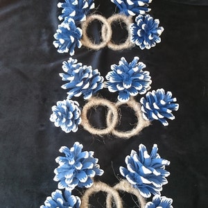 May include: Three sets of blue and white painted pine cones tied together with twine to create napkin rings.
