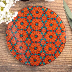 May include: Round glass cutting board with a vibrant floral pattern in orange and teal. The design features a repeating pattern of stylized flowers. The board is placed on a wooden surface, with a knife and other kitchen items nearby.