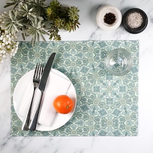 May include: A green and blue patterned placemat with a white plate, a fork, a knife, a napkin, and an orange on top.