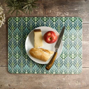 May include: A rectangular placemat featuring a repeating geometric leaf pattern in shades of teal, green, and light blue. A white plate sits on the placemat, holding a slice of hard cheese, a red apple, and a piece of bread with a metal knife.