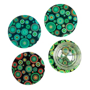 Può includere: Quattro sottobicchieri rotondi con un design a cerchi concentrici colorati. I sottobicchieri presentano uno sfondo scuro con cerchi verdi, turchese, arancioni e gialli di varie dimensioni. Un bicchiere trasparente è appoggiato su uno di essi.