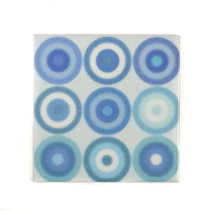 May include: Square ceramic tile featuring a repeating pattern of nine concentric circles in shades of blue and white. The circles vary in size and color, creating a retro, geometric design. The tile has a glossy finish and a light blue background.