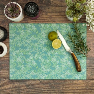 May include: A rectangular cutting board with a floral pattern in shades of green and white. A lime cut in half, a knife, and sprigs of rosemary are on the board. The board is on a wooden surface.