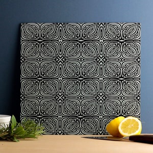 May include: A square tile with a black and white geometric pattern. The pattern is made up of repeating shapes that resemble leaves or flowers.