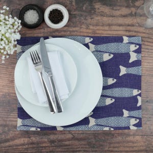 May include: A rectangular placemat featuring a dark blue background with a repeating pattern of light blue fish. A large white dinner plate is topped with a smaller white salad plate, a folded white napkin, a silver fork, and a silver knife.