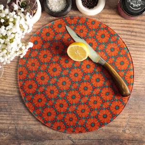 May include: A round orange and teal glass chopping board with a floral pattern. A knife and a lemon wedge are on the board.