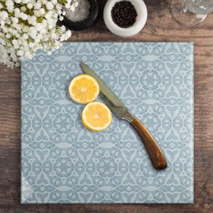 May include: A square, pale blue chopping board with a geometric design, a knife with a wooden handle, and two lemon halves. The board is surrounded by small bowls of salt and pepper, and a bouquet of white flowers.