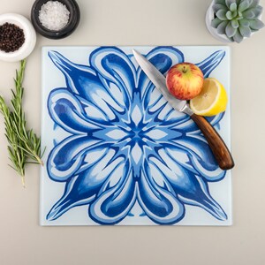 May include: Square glass cutting board with a blue floral design. A stainless steel knife rests on the board next to a red and yellow apple and a lemon half. Rosemary, salt, pepper, and a succulent are also present.