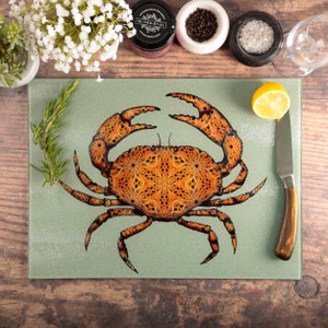 May include: A green glass cutting board with a detailed illustration of a crab with an orange and brown mandala pattern. The crab is centered on the board and has its claws raised.