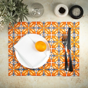 May include: A rectangular placemat featuring a repeating orange, white, and dark blue floral tile pattern. A white plate holds a folded white napkin and a single orange fruit. A silver fork and a silver knife are placed to the right of the plate.