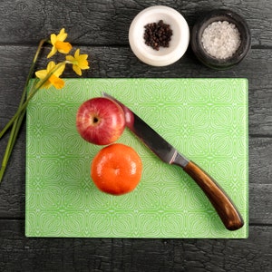 May include: A green chopping board with a decorative pattern, featuring a red apple, an orange, and a knife with a wooden handle.