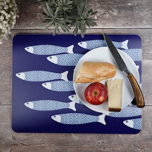Op de afbeelding: Een rechthoekige placemat met een donkerblauwe achtergrond en een herhalend patroon van witte vissen met ingewikkelde blauwe geometrische schubben. Op een wit bord liggen een stuk stokbrood, een rode appel en een plakje kaas met een mes.