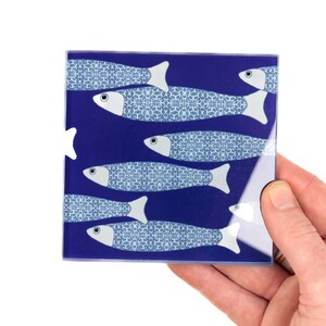 May include: A blue glass coaster featuring a design of white fish with blue and white patterned scales on a dark blue background.