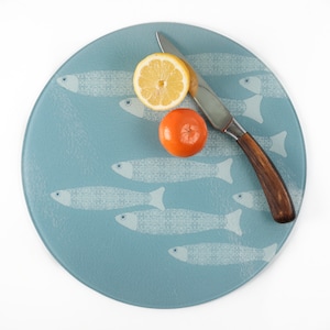 May include: A blue glass chopping board with a white fish design. There is a lemon and an orange on the board, along with a silver knife.