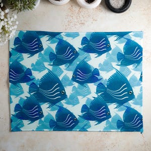 Op de afbeelding: Een witte placemat met een blauw en wit vispatroon. De vissen zijn gestileerd en hebben een blauw en wit gestreept patroon.
