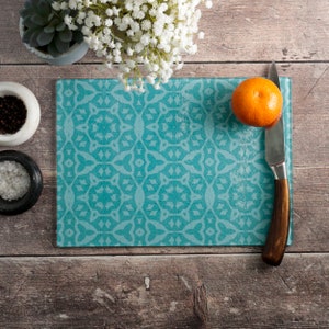 Puede incluir: Una tabla de cortar de vidrio turquesa con un patrón decorativo. Un cuchillo y una naranja están sobre la tabla.