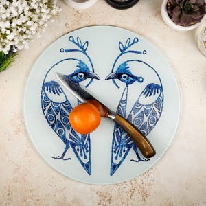 May include: A round glass cutting board with a blue and white design of two stylized birds facing each other. A small orange sits on the board, and a silver knife is placed between the birds.