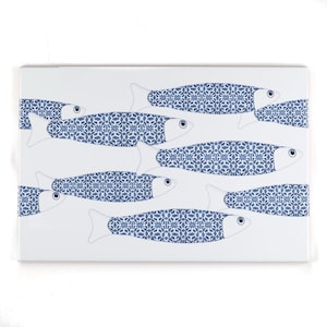 May include: A rectangular white ceramic tile featuring a pattern of stylized blue fish. Each fish has a detailed blue and white floral design on its body, with white fins and a single black eye. The fish are arranged in a repeating pattern.