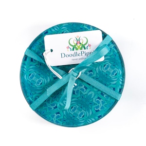 May include: A set of teal coasters with a floral design, tied with a teal ribbon. A white tag with the text "DoodlePipp" and "never ordinary" is attached. The coasters have a diameter of approximately 10 cm.