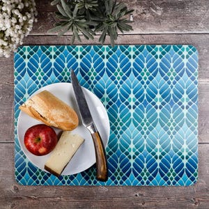 May include: A rectangular placemat featuring a repeating blue and teal leaf-like geometric pattern. A white plate sits on top, holding a piece of crusty bread, a red apple, a wedge of cheese, and a metal knife with a dark wooden handle.