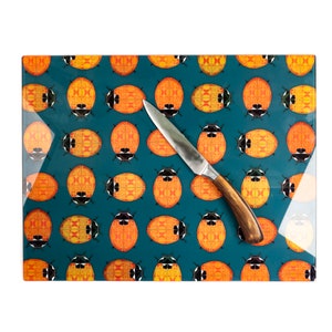 May include: A rectangular glass chopping board with a teal background and an orange ladybug pattern. A stainless steel knife with a wooden handle is on the board. The ladybugs have black and yellow details.