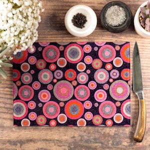 May include: A black cutting board with a pink and purple geometric pattern of overlapping circles. The cutting board is made of glass and has a shiny finish.