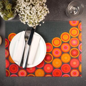 May include: A gray placemat with a colorful pattern of overlapping circles in shades of orange, red, and pink. A white plate with a white napkin and silver cutlery is on top of the placemat.