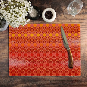 May include: A rectangular glass chopping board featuring a repeating pattern of orange and yellow circles and shapes. A stainless steel knife with a wooden handle is on the board. The board is on a wooden surface.