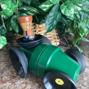 Könnte beinhalten: Ein skurriles, handgefertigtes Gartenkunstwerk. Ein grün bemalter Terrakottatopf bildet die Karosserie des Autos, mit schwarzen Rädern. Eine kleine, lächelnde Terrakotta-Topffigur mit blauen Latzhosen sitzt in einer schwarzen Schüssel.