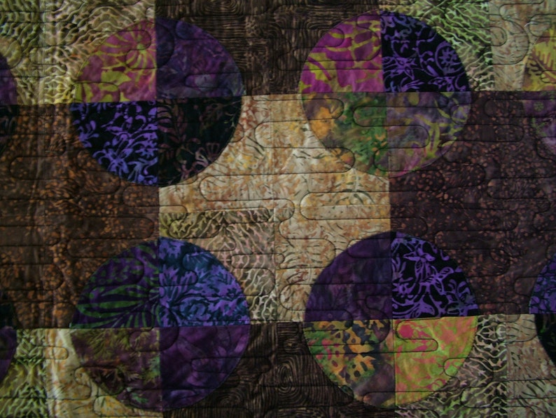 K&ouml;nnte beinhalten: Eine Steppdecke mit Patchwork-Muster, die Kreise in verschiedenen Schattierungen von Lila, Gr&uuml;n und Braun zeigt. Die Kreise sind in einem Gittermuster angeordnet und mit einem wellenf&ouml;rmigen Muster gesteppt.