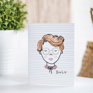 May include: A greeting card with a cartoon illustration of a person with red hair wearing glasses and a pink bow. The card has a gray and white striped background and the text "Barb" with a pink heart.