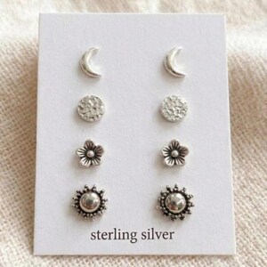 May include: A set of four pairs of sterling silver stud earrings on a white card. The earrings include crescent moons, textured circles, flower shapes, and sunburst designs. The card has the words "sterling silver" printed at the bottom.