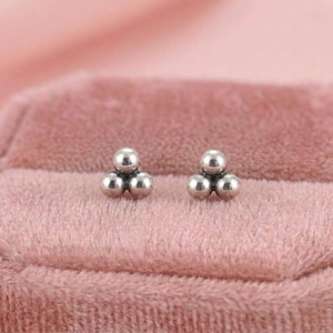 May include: A pair of silver stud earrings, each with three small, spherical beads in a triangular cluster. The earrings are presented on a soft, pink velvet surface, creating a delicate and elegant presentation.