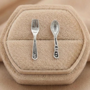 May include: Miniature silver fork and spoon set. The fork has three tines and a decorative handle. The spoon has a rounded bowl and a handle with a decorative design. The set is displayed on a beige surface.