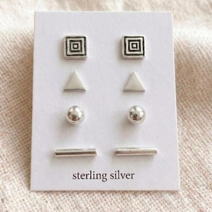 May include: A set of four pairs of sterling silver stud earrings on a white card. The earrings include square, triangle, ball, and bar shapes. The card has the words "sterling silver" printed at the bottom.