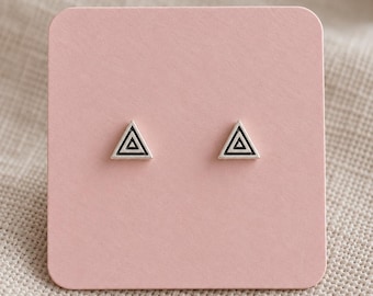 Puede incluir: Un par de pendientes de botón con forma de triángulo, cada uno con un recorte triangular más pequeño en el centro. Los pendientes están montados sobre una tarjeta de presentación cuadrada de color rosa claro con esquinas redondeadas.