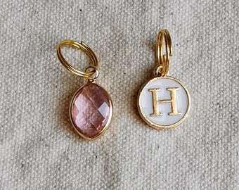 May include: Two gold keychains with charms. One charm is a pink oval-shaped gemstone. The other charm is a white circle with the letter "H" in gold.