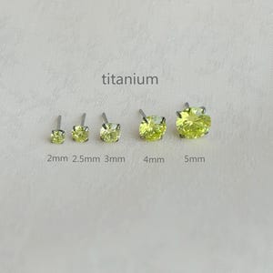 May include: Five titanium stud earrings with light green gemstones, ranging in size from 2mm to 5mm. The word "titanium" is printed above the earrings. The earrings are displayed on a white surface.