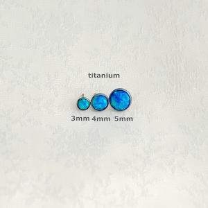 Peut inclure: Trois boucles d'oreilles en titane opale bleue de tailles 3 mm, 4 mm et 5 mm. Les boucles d'oreilles sont rondes et d'un bleu vif et irisé. Le mot "titanium" est au-dessus des boucles d'oreilles.
