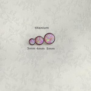 May include: Three titanium stud earrings with pink opal-like centers, sized 3mm, 4mm, and 5mm. The word "titanium" is above the earrings. The earrings are arranged in a row on a white textured surface.