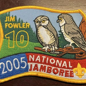 May include: A yellow, red, blue, and white embroidered patch with the text "Jim Fowler 10 2005 National Jamboree" and two owls perched on a branch. The patch features a scout's fleur-de-lis.