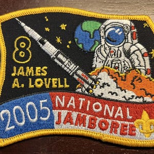 May include: Embroidered patch with a black background, featuring an astronaut, rocket, and Earth. Text includes "8 JAMES A. LOVELL" and "2005 NATIONAL JAMBOREE". The patch has a yellow border and a Boy Scout emblem.